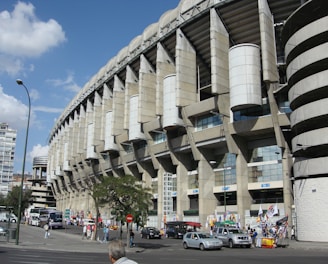 A large, modern sports stadium with a curved roof and prominent architectural features dominates the scene. Surrounding the structure, people and several vehicles are visible, indicating a busy urban environment. Flags and banners suggest an event or gathering. Nearby, urban buildings and trees add to the bustling atmosphere.